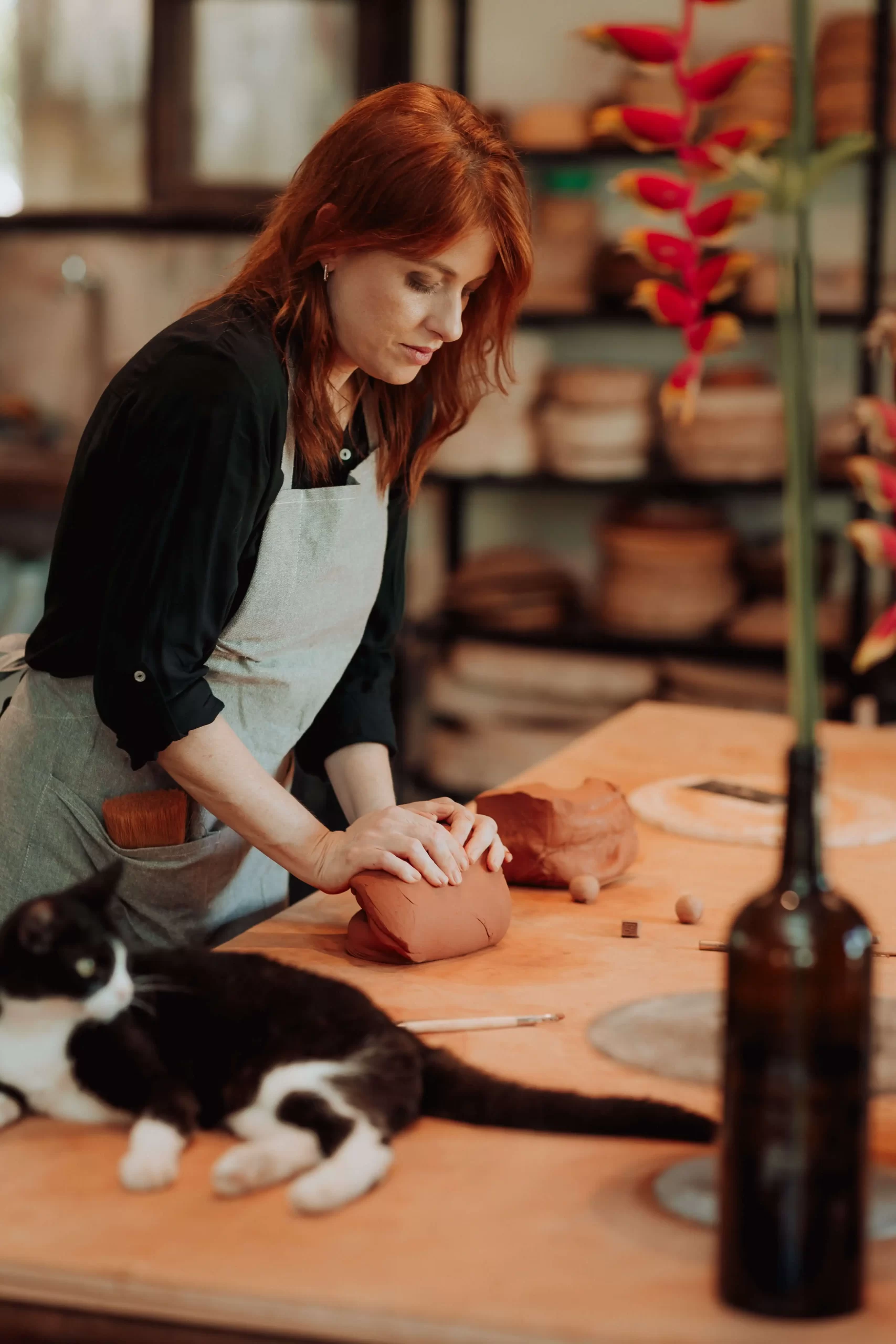 Vívian Vieira preparando argila em ateliê sensorial da Bone Ceramics, com gato deitado sobre a mesa e ambiente criativo ao fundo