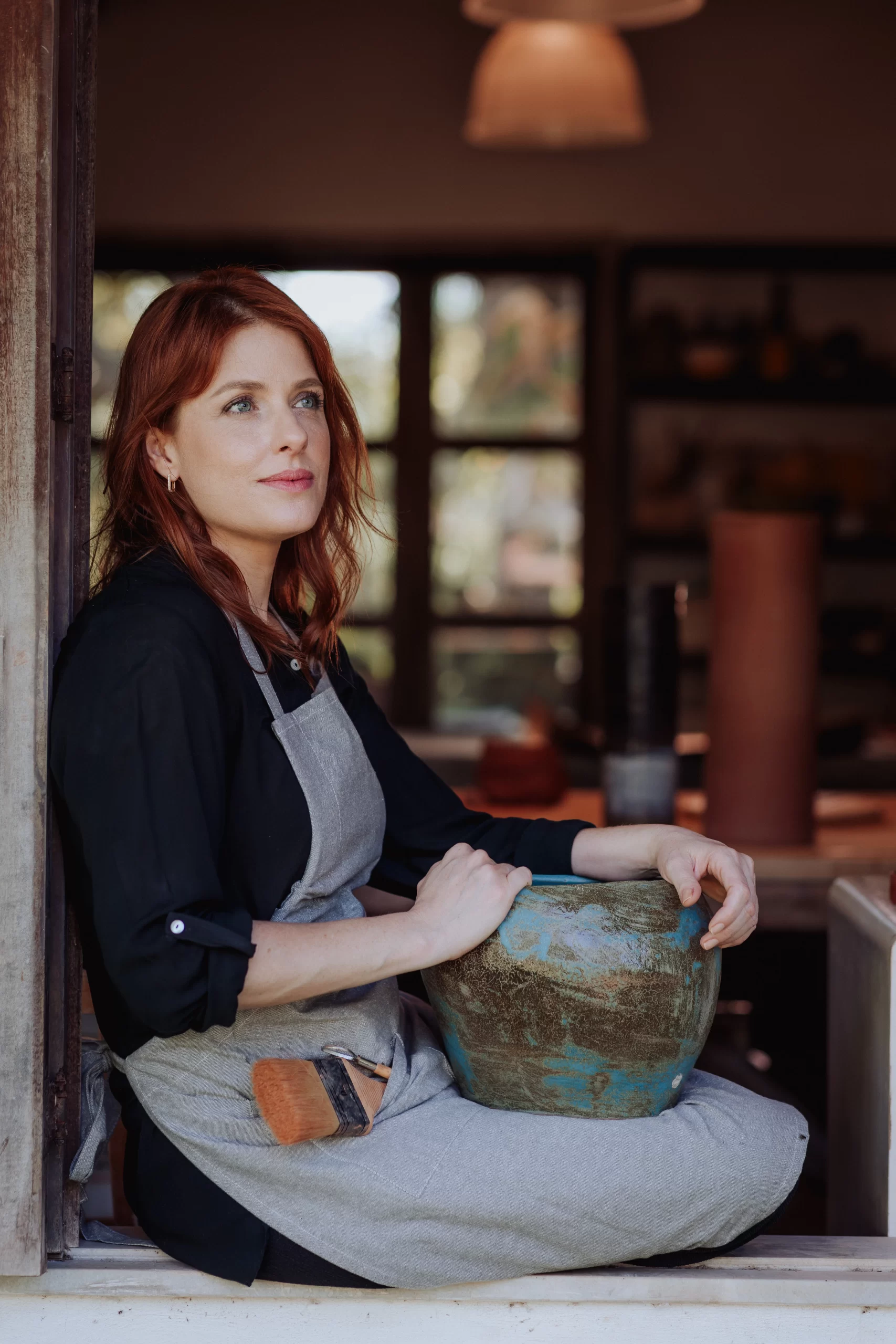 Artista Vívian Vieira sentada à janela com uma peça de cerâmica azul-esverdeada no colo, em momento contemplativo no ateliê da Bone Ceramics